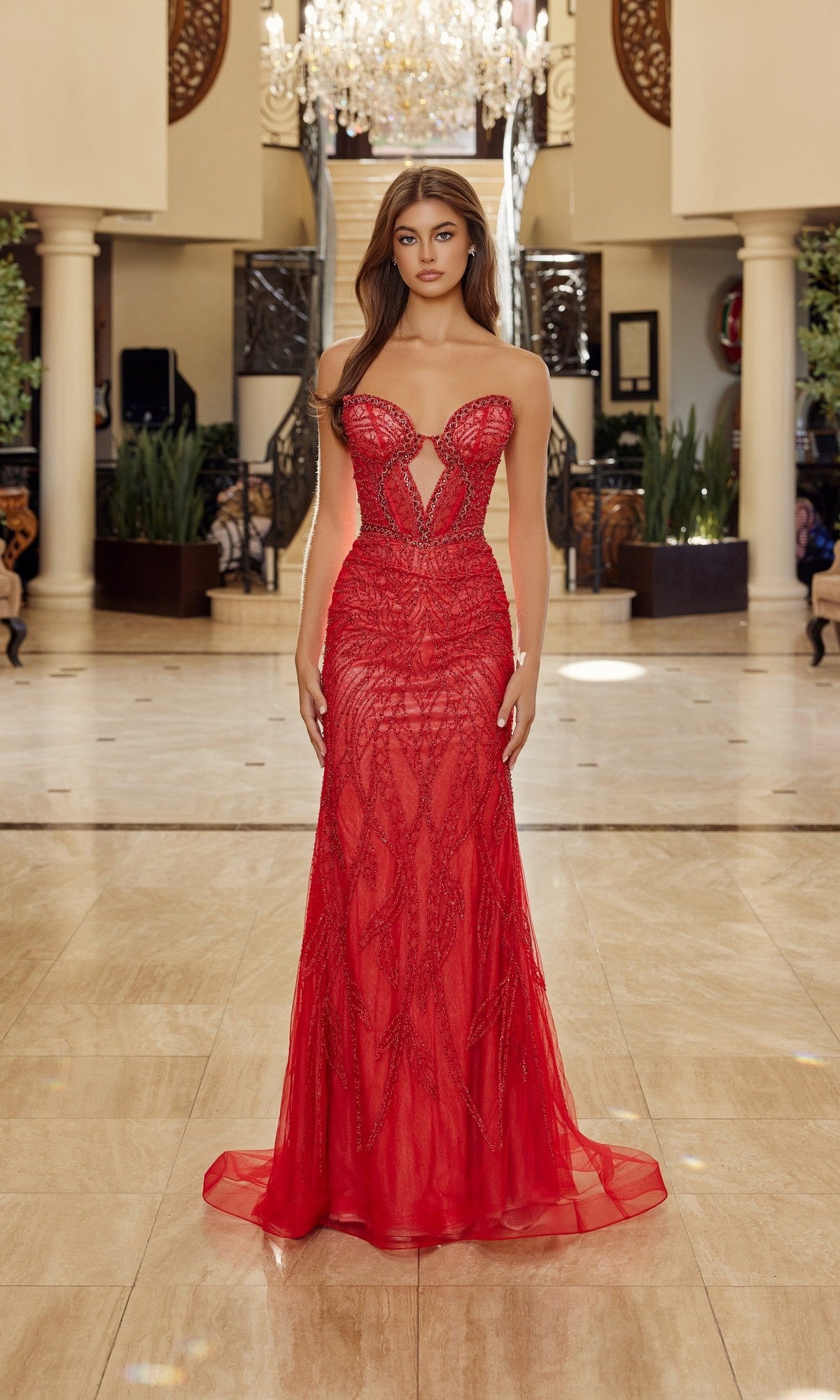 A woman stands in a spacious, elegant foyer wearing the Nox Anabel C1848 long prom dress in red/nude, featuring a strapless romantic design with intricate beadwork and a flattering sweetheart neckline.